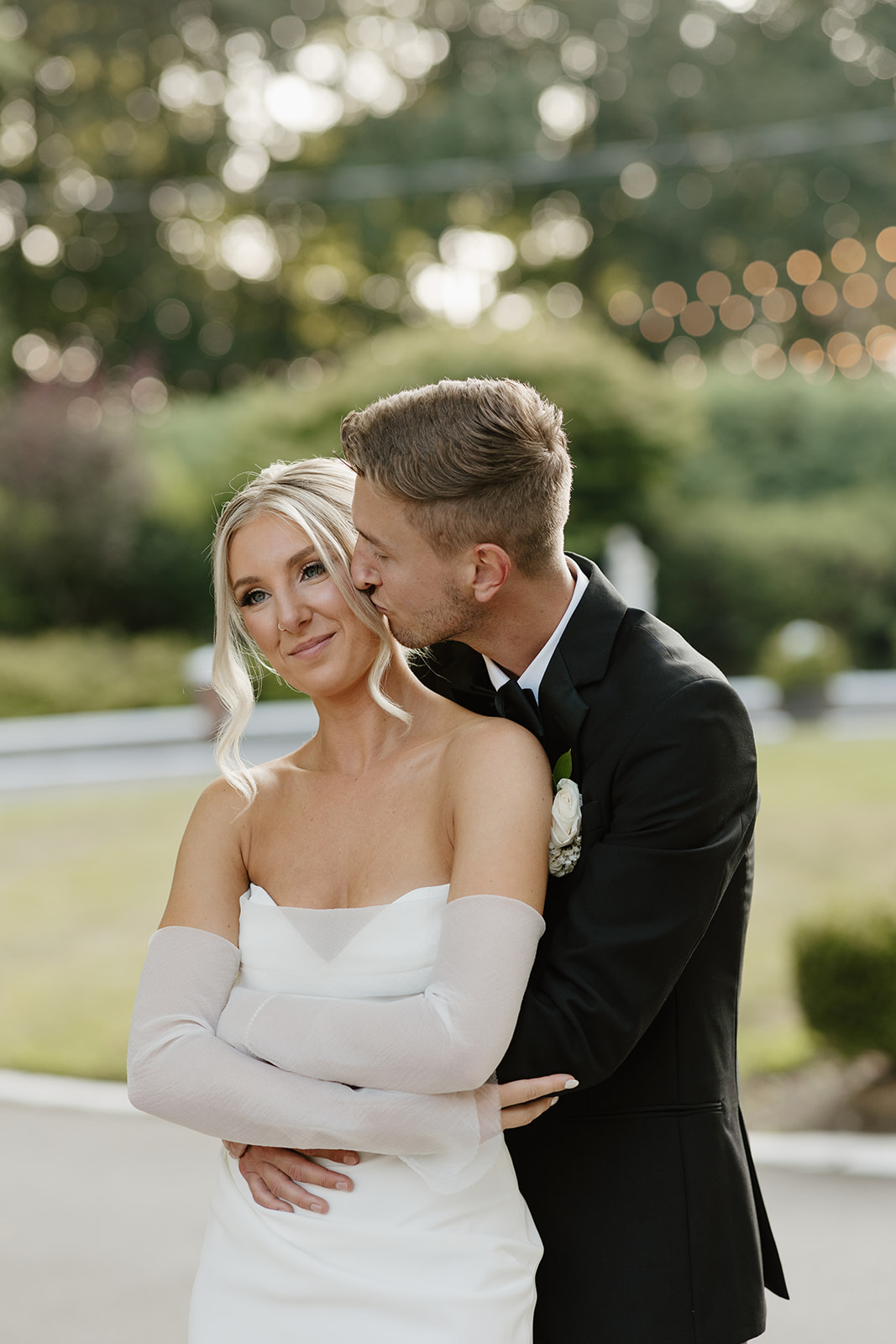 Groom kisses the bride's cheek while hugging her from behind, set against a soft, green background.